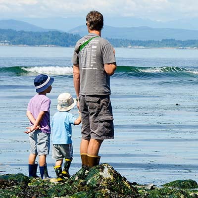 An father standing on a beach their two young children as all three look at waves coming in to shore.